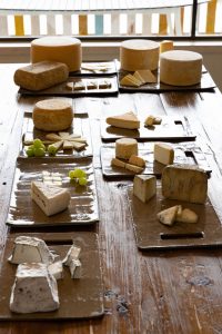 סוגים שונים של גבינות מוצגים על שולחן עץ, כל אחד על לוח נפרד. חלק מהצלחות כוללות גם אשכולות קטנים של ענבים ירוקים. הגבינה היא בצורות, גדלים ומרקמים שונים, מה שמצביע על מבחר מגוון.
