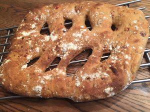 כיכר לחם מלבנית וכפרית עם קרום חום-זהוב וכמה חרכים גדולים מתקררת על מתלה מתכת המונח על שולחן עץ. משטח הלחם מאובק קלות בקמח.