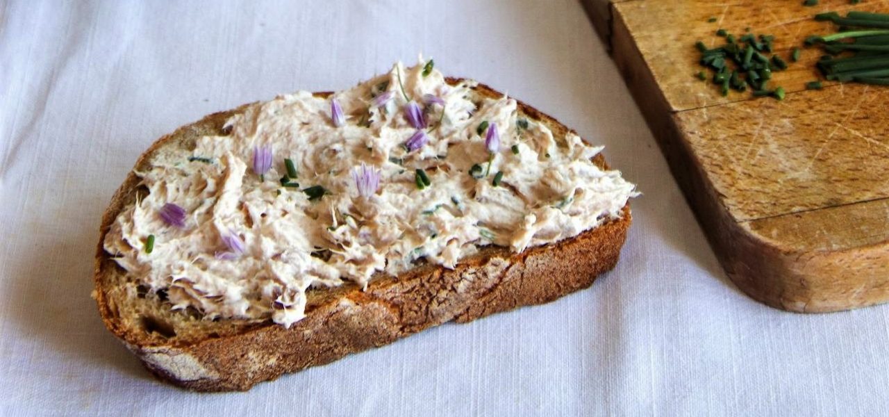 צנצנת דג מרוחה בכף לצד פרוסת לחם שעליה ממרח. קרש חיתוך מעץ עם עירית ופרחי עירית, לצד פרוסות לחם נוספות. הכל מונח על מפה לבנה.