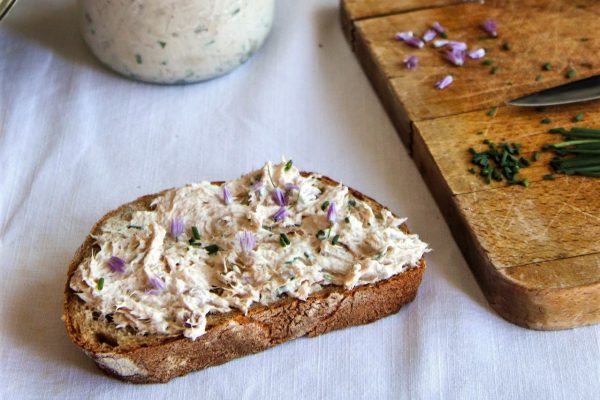 צנצנת דג מרוחה בכף לצד פרוסת לחם שעליה ממרח. קרש חיתוך מעץ עם עירית ופרחי עירית, לצד פרוסות לחם נוספות. הכל מונח על מפה לבנה.