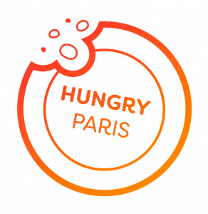 לוגו עיגולי עם קו מתאר כתום בגוון מדורג, סימן נשיכה בחלק העליון השמאלי, והמילים HUNGRY PARIS במרכז. הרקע שקוף.