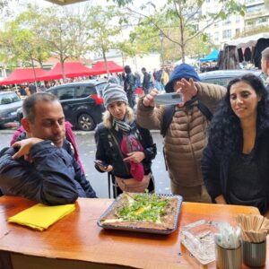 אנשים עומדים בדוכן אוכל חיצוני, כאשר שניים מהם מצלמים מנה מלבנית. ברקע ניתן לראות עצים, מכוניות חונות ואוהלים אדומים באווירה סוונת.
