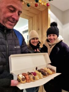 גבר מחזיק קופסה פתוחה עם מבחר מאפים, בעוד שתי נשים לבושות בבגדי חורף עומדות לידו ומחייכות. הם נמצאים בתוך המבנה, ליד פתח עץ מקושט באורות שרשרת אוקישוטים.