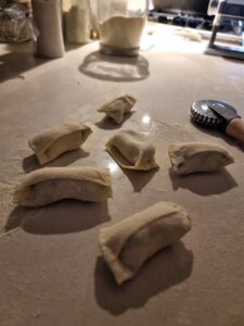 שבע חתיכות של בצק חי ומקופל מונחות על משטח מקומח, כשברקע גלגלת בצק וצנצנות מרכיבים, מה שמרמז על הכנת פסטה או כיסונים תוצרת בית.