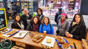 קבוצת שבעה מבוגרים עומדת מסביב לדלפק עץ עם לחם, גבינות ומאפים בשוק. ברקע נראות מדפי סחורה ודוכן בשרים. רוב האנשים לובשים מעילים.