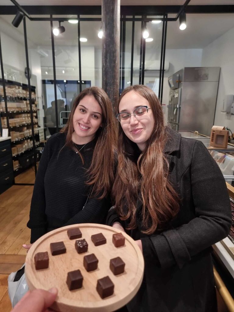 שתי נשים לבושות כהה עומדות בחלל פנימי ליד דלפק עץ. בקדמה, יד מחזיקה מגש עגול מעץ שעליו כמה קוביות שוקולד מרובע. ברקע נראות מדפים וביקורות זכוכית.