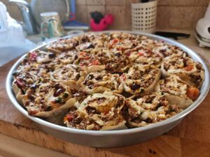 תבנית אפייה עגולה ממתכת מלאה במאפים בצורת ספירלה, המכילים תערובת של בשר טחון, ירקות וגבינה, מונחת על קרש חיתוך עץ במטבח.