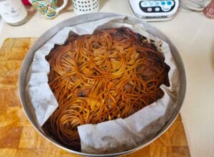 תבנית אפייה עגולה ממתכת מרופדת בנייר אפייה מכילה ספגטי אפוי עם שכבה עליונה זהובה-חומה, מונחת על קרש חיתוך מעץ על משטח במטבח. פריטים שונים של מטבח נראים ברקע.