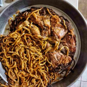 תבנית מתכת המכילה אטריות מבושלות שמעורבבות עם חתיכות בשר צלוי, חלקן מסודרות בנתחים גדולים בצד אחד, בעוד שהאטריות הנותרות מפוזרות מסביב.