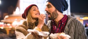 אישה עם כובע צמר אדום מחייכת בזמן שהיא מאכילה גבר שלובש כובע כחול וחולצה משובצת. שניים לבושים חמים, עומדים בחוץ בלילה וחטיפים בידיהם.