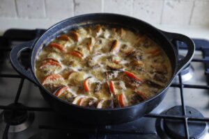 מחבת שחורה על הכיריים מכילה תבשיל אפוי עם עגבניות פרוסות ובשר השחום ברוטבנת, מסודרים בתבנית שמגלית. התערובת מבעבעת ונראית מבושלת טרי.