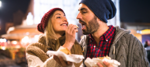 אישה עם כובע צמר אדום וצעיף מאכילה גבר עם כובע צמר כחול וחולצה משובצת. שניהם אוכל ומחייכים, עומדים בחוץ מואר מאוד.